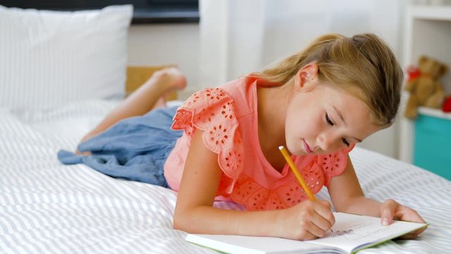 people, children, creativity and art concept - little girl lying on bed and drawing something in sketchbook with graphite pencil at home