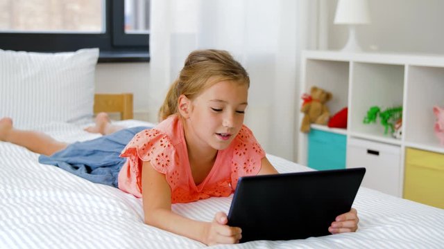 children, technology and communication concept - smiling girl having vide call on tablet computer lying on bed at home