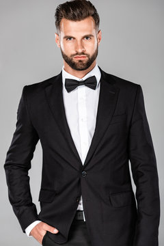 elegant man in black suit and bow tie with hand in pocket isolated on grey