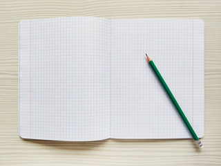 White school notebook in a cage is on a wooden table. Green pencil rests on a checkered exercise book without notes. Empty notepad is on a desk. Square texture