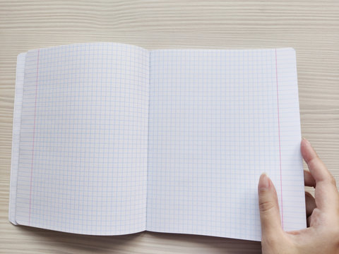 White open school checkered exercise book in a cage is on a wooden table. White woman opens a notebook without notes. The right hand of a white European woman holding a empty notepad with square