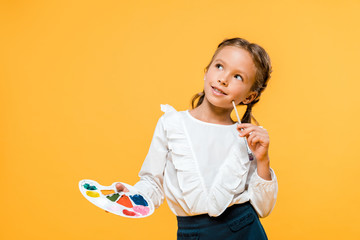 pensive schoolkid holding palette and paintbrush isolated on orange