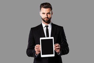 handsome businessman in black suit showing digital tablet with blank screen isolated on grey