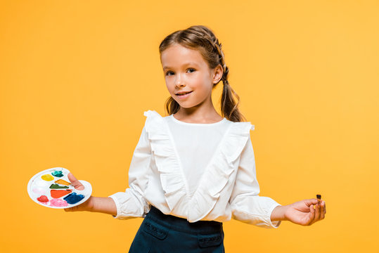 cheerful schoolgirl holding palette and paintbrush isolated on orange - Powered by Adobe
