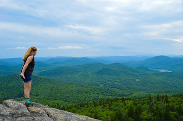 A female hiker cautiously leans over the edge of a mountain, looking down with the Adirondack mountain ranges on display in the distance.