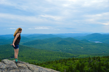 Naklejka na ściany i meble A female hiker cautiously leans over the edge of a mountain, looking down with the Adirondack mountain ranges on display in the distance.