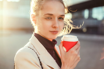 A beautiful girl in a beige jacket drinks coffee from a glass talking on the phone, wearing sunglasses works in the office in the business center, the manager, the employee of the company, happy face