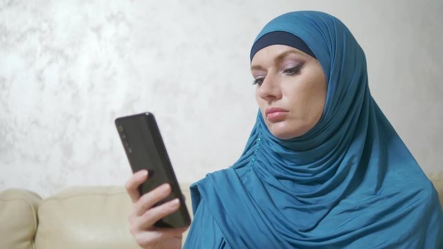 beautiful muslim woman in blue hijab using a mobile phone sitting on a sofa