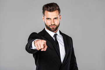 successful businessman in black suit pointing with finger at camera isolated on grey