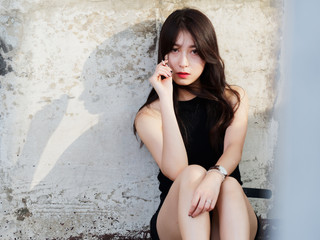 Beautiful young woman with long black hair sitting on floor, leaning against wall in sunny afternoon, looking at camera, holding a lighted cigarette in her hand, smoking silhouette shadows on wall.