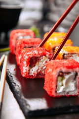 Chopstick holding chicken maki sushi rolls with rice, cream cheese, chicken fillet, tomato, cucumber, red flying fish caviar and nori. Fresh tory teriyaki uramaki roll on black stone background © Ryzhkov
