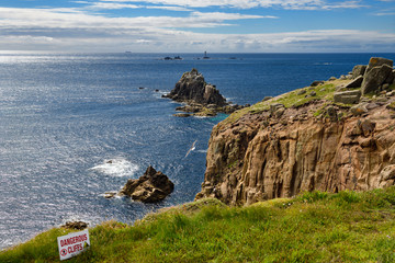 Sign for dangerous cliffs at the edge of Land's End looking to Armed Knight and Longships Lighthouse islands in the Atlantic Ocean Cornwall England