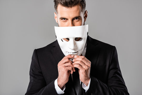 Sly Handsome Businessman In Black Suit Taking Off White Mask Isolated On Grey