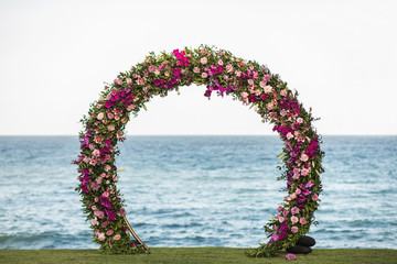 Wedding round arch decorated with beautiful colorful orchid and rose flowers on the beach. Ocean on...