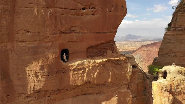 Africa religion and natural scenery - aerial drone shot of Orthodox priest at remote rock-hewn monolithic monastery (Abuna Yemata Guh), reveals spectacular surreal desert mountain landscape in Ethiopi