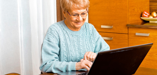 Smiling senior woman, grandmother sitting in home and working on laptop.