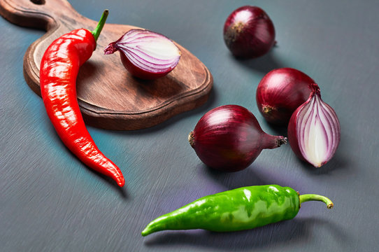 Red and green chilli peppers near purple onion and wooden cutting board on dark concrete background