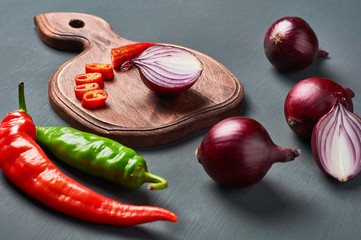 Red and green chilli peppers near purple onion and wooden cutting board on dark concrete background