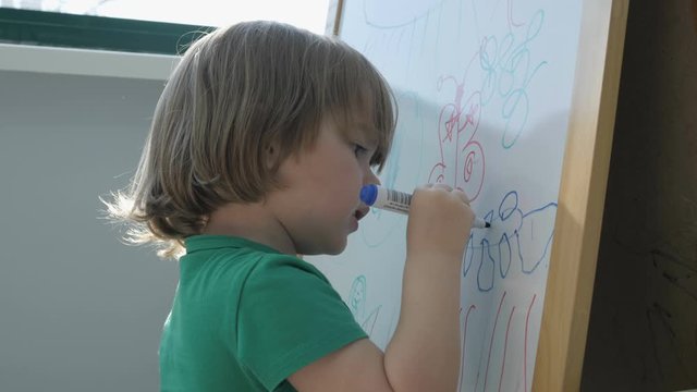 Child painting on easel. Closeup. Cute little boy is drawing with pencils in preschool. Child draw with colorful markers and smile.