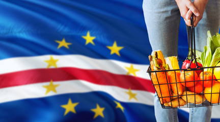 Woman is holding supermarket basket, Cape Verde waving flag background. Economy concept for fresh fruits and vegetables.