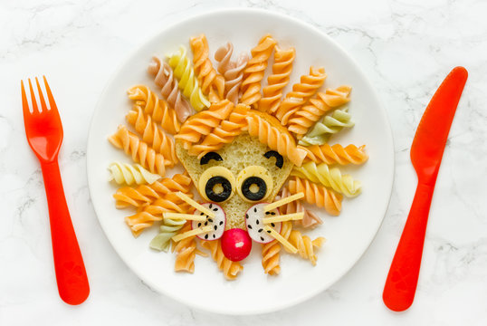 Fun food idea for kids lunch, animal shaped food art - colorful fusilli vegetables pasta with sandwich like a cute lion head on white plate top view