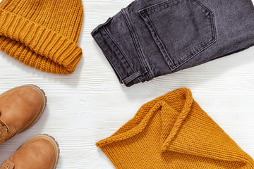 Bright fashion clothes on white wood with copy space. Knitted scarf and cap orange colored, comfort leather shoes and Black jeans. Flat lay with warm womens clothing. Top view.