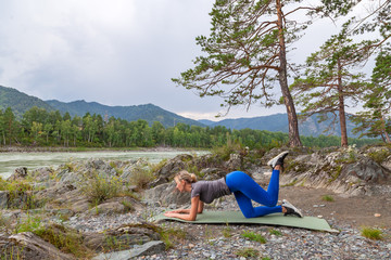 Slim young healthy sports girl doing the donkey kick exercise on all fours arching back straightening leg up concept sport, fitness, lifestyle. Work out and warm up outdoors in mountains near river.