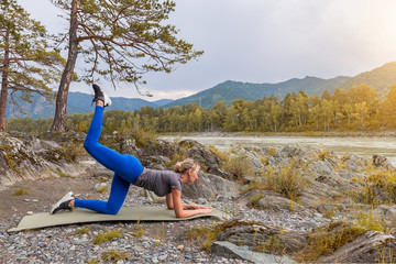 Slim young healthy sports girl doing the donkey kick exercise on all fours arching back straightening leg up concept sport, fitness, lifestyle. Work out and warm up outdoors in mountains near river.