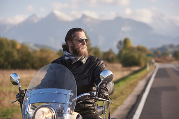 Portrait of handsome bearded biker in black leather jacket holding motorcycle handles on country roadside on blurred background of green landscape, distant white mountain peaks and bright sky.