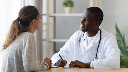 Woman talking to smiling african doctor noting at medical consultation