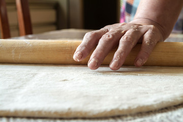 Senior woman hands are rolling out dough in flour with rolling pin in her home kitchen. Homemade noodle or pasta production by grandma. Closeup, selective focus