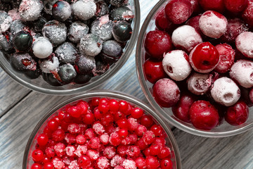 Ripe frozen sweet cherries, red currant and black currant with hoarfrost in the transparent glass cups on blue wooden background. Natural organic healthy food. Closeup, selective focus, top view