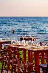 Tables in outdoor cafe or restaurant served for dinner on crowded beach with sea view, summer holidays