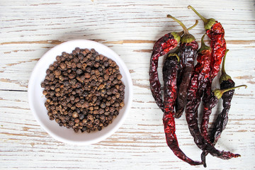 Black peppercorns on small white plate on white wooden table background with dried black chilli peppers,top view,space for text,spices
