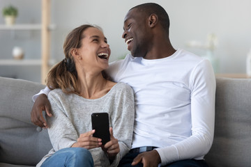 Cheerful interracial couple laughing holding phone sit together on couch