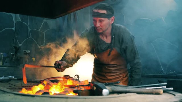 Male blacksmith uses a metal poker to move coal. Blacksmith forging iron in workshop.