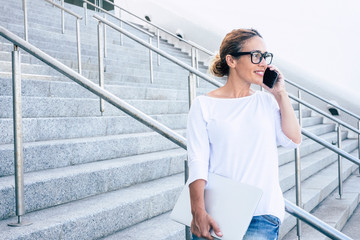 business woman with computer or laptop under her arm is calling and talking with her phone or cell about working looking in front of her on stairs in commercial district