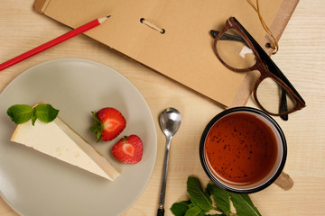 An open notebook with a red pencil and glasses lies next to a plate with a cheesecake and a cup of tea on a light wooden table.