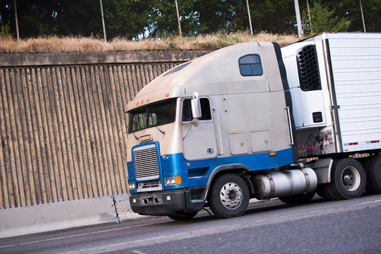 Old cab-over big rig semi truck transporting food cargo in refrigerator semi trailer moving on the road with concrete wall on the side