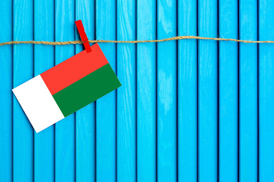 Flag of Madagascar hanging on clothesline attached with wooden clothespins on aqua blue wooden background. National day concept. - Powered by Adobe
