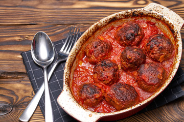 Meatballs cooked with tomato sauce in white casserole on wooden rustic table, with spoon and fork. black napkin with white cage. top view