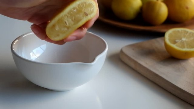 Squeezing fresh lemon juice by hand into white ceramic spouted bowl, letting pips and seeds fall in. close up.