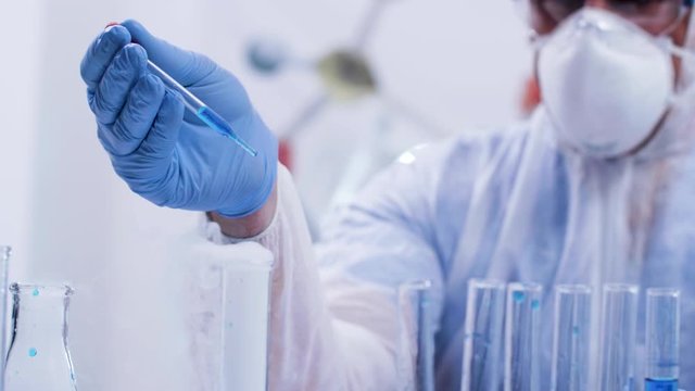 Scientist in modern laboratory with coverall protection equipment pouring blue reagent using pipette. Scientific process.