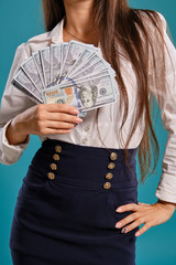 Brunette girl in glasses, wearing in a black short skirt and white blouse is posing holding a fan of hundred dollar bills against a blue background.