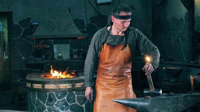 Male forger stands near an anvil, holding a hammer. Blacksmith forging iron in workshop.