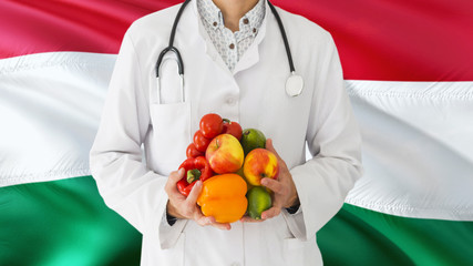 Doctor is holding fruits and vegetables in hands with Hungary flag background. National healthcare concept, medical theme.