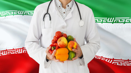 Doctor is holding fruits and vegetables in hands with Iran flag background. National healthcare concept, medical theme.