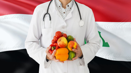 Doctor is holding fruits and vegetables in hands with Iraq flag background. National healthcare concept, medical theme.