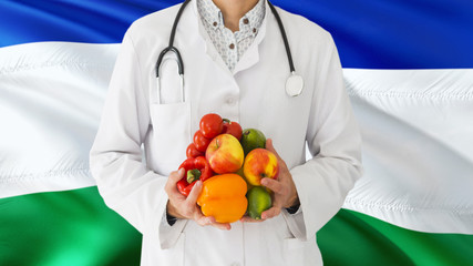 Doctor is holding fruits and vegetables in hands with Lesotho flag background. National healthcare concept, medical theme.