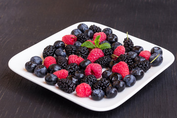 Berries: raspberries and blueberries, blackberries on a white plate, black background.